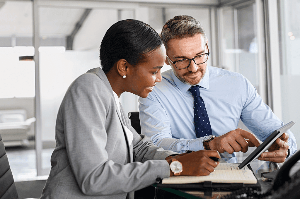 Two professionals discussing work at a desk. One person writes on a notepad while the other gestures at a tablet, both smiling.