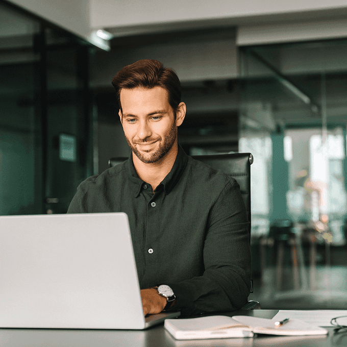 Smiling man in a dark shirt sits at a desk, working on a laptop in a modern office with glass walls.